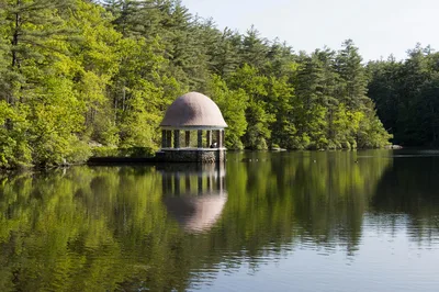 Tranquil scene of a gazebo reflecting in a pond surrounded by lush trees in Fitchburg, MA.